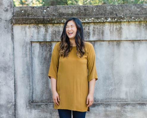woman wearing a modern, mustard yellow tunic standing in front of a moss covered concrete wall
